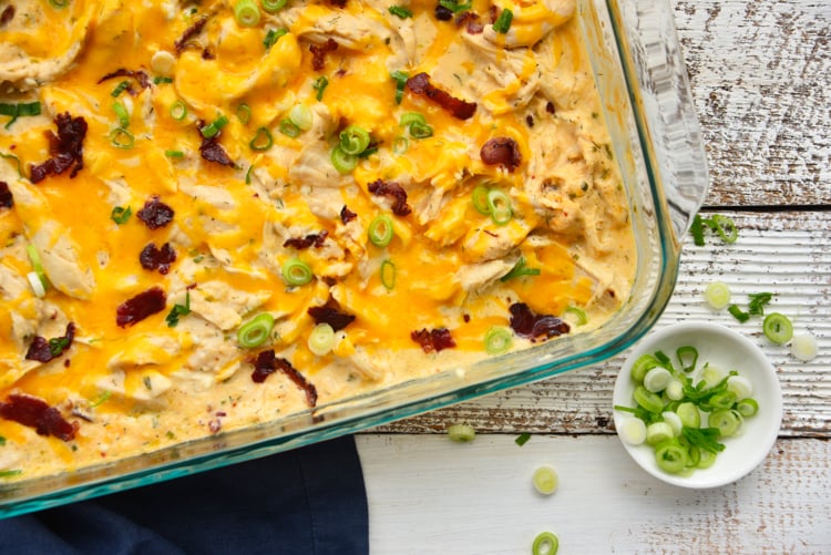 fully baked chicken topped with shredded cheddar cheese, bacon, and scallions in a glass baking dish