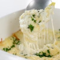 square image of Baked Creamy garlic parmesan hot artichoke dip in a white square casserole dish with a silver spoon taking a scoop