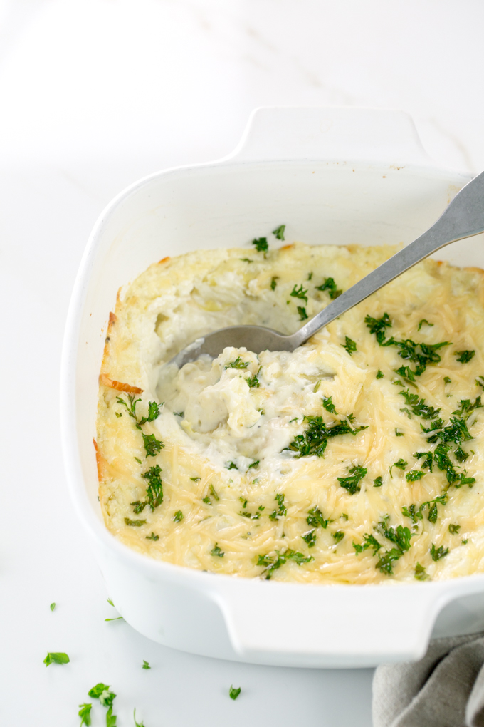 Vertical image of Baked Creamy garlic parmesan hot artichoke dip in a white square casserole dish with a spoon