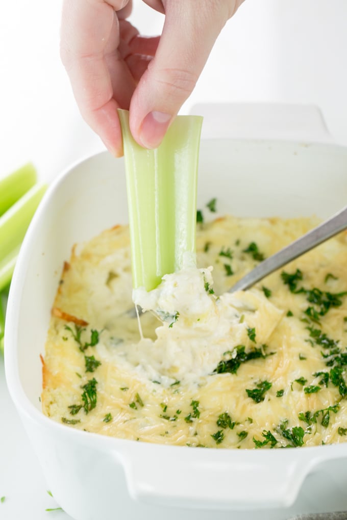Vertical image of Baked Creamy garlic parmesan hot artichoke dip in a white square casserole dish with celery