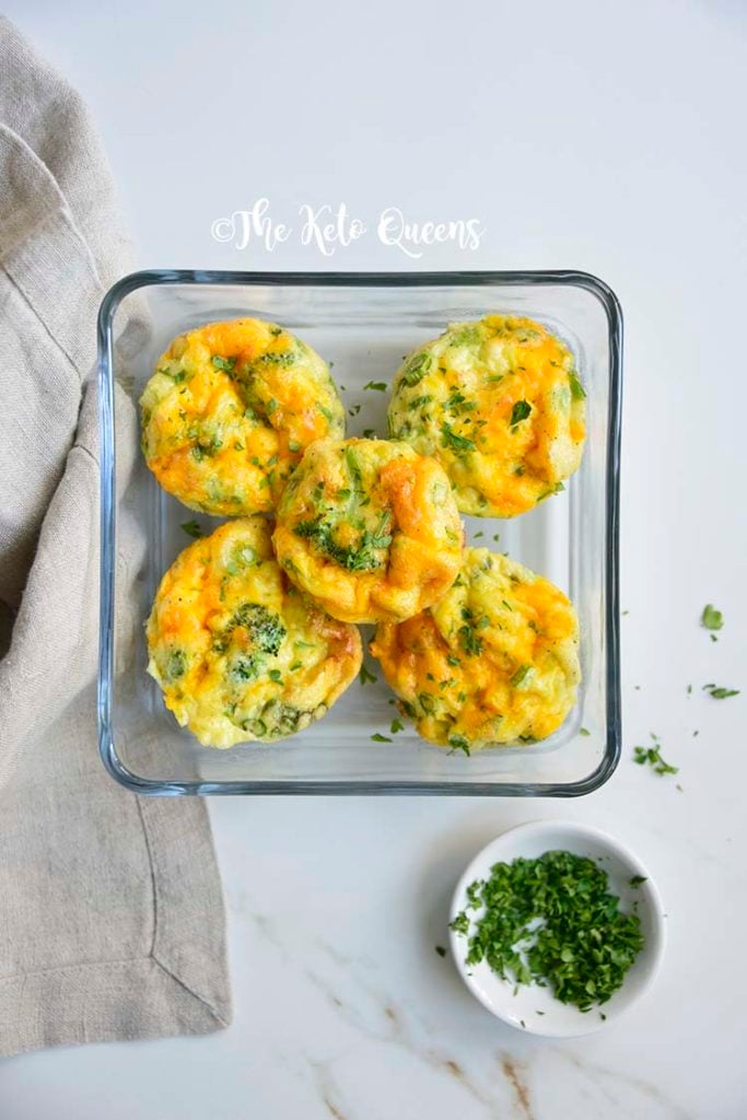 Overhead close up vertical image of broccoli and cheddar egg muffins in a glass storage container on a white background with parsley