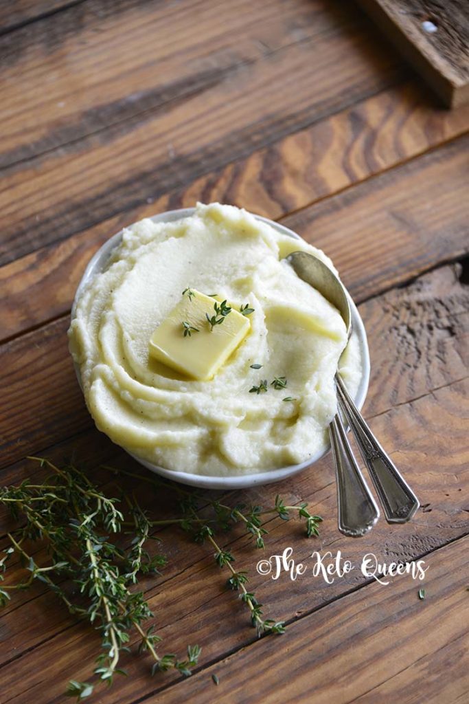 Pinterst image for low carb butter garlic mashed cauliflower Vertical image of mashed cauliflower with butter and fresh time in a bowl with 2 spoons