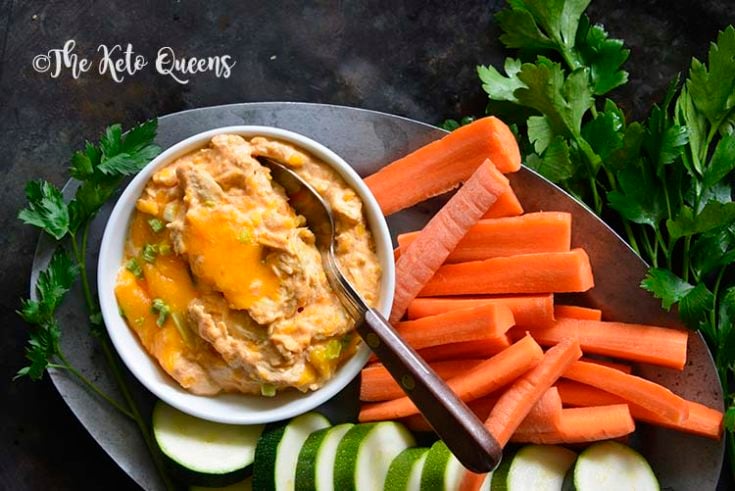 This Super Easy Buffalo Chicken Dip Recipe is the perfect appetizer for football Sundays, tailgating parties or to just have on hand as a keto snack. horizontal image of buffalo chicken dip in a small bowl with a spoon, with carrots, cucumber and cilantro