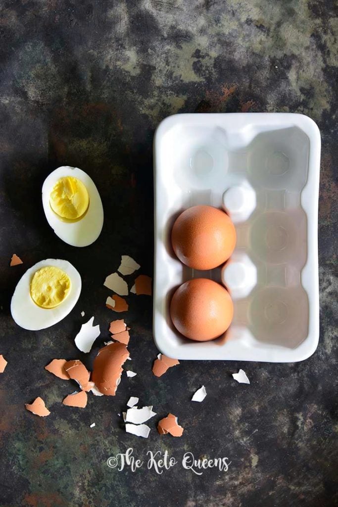 a vertical overhead image of 2 brown leftover hard boiled eggs in a carton and 1 hard boiled egg split in half on a dark background