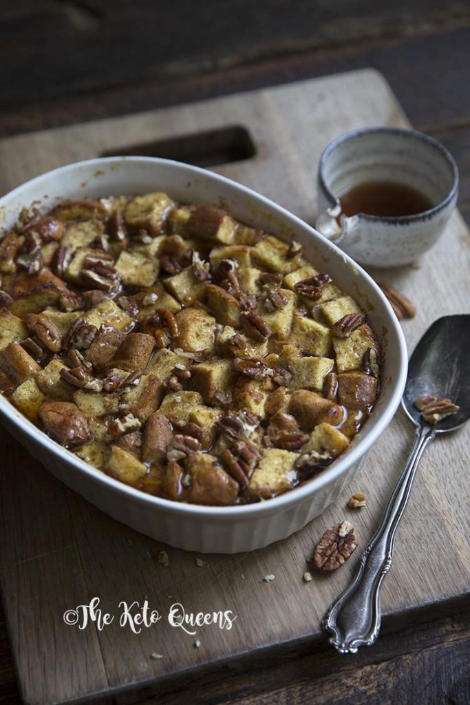 zoomed in vertical image of maple pecan french toast casserole in a white casserole dish, with a spoon and low carb syrup on a wood background