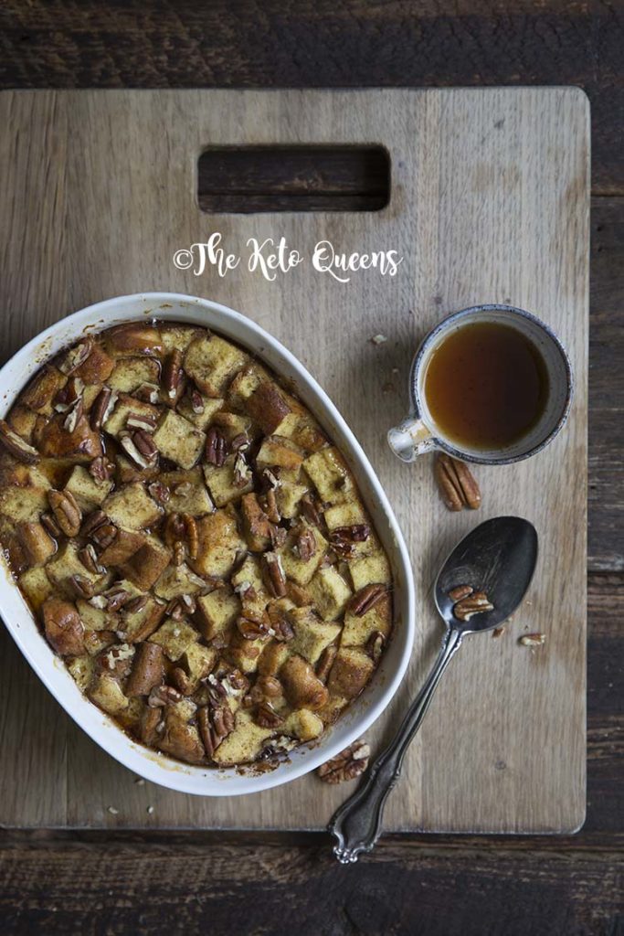 overhead vertical image of maple pecan french toast casserole in a white casserole dish, with a spoon and low carb syrup on a wood background