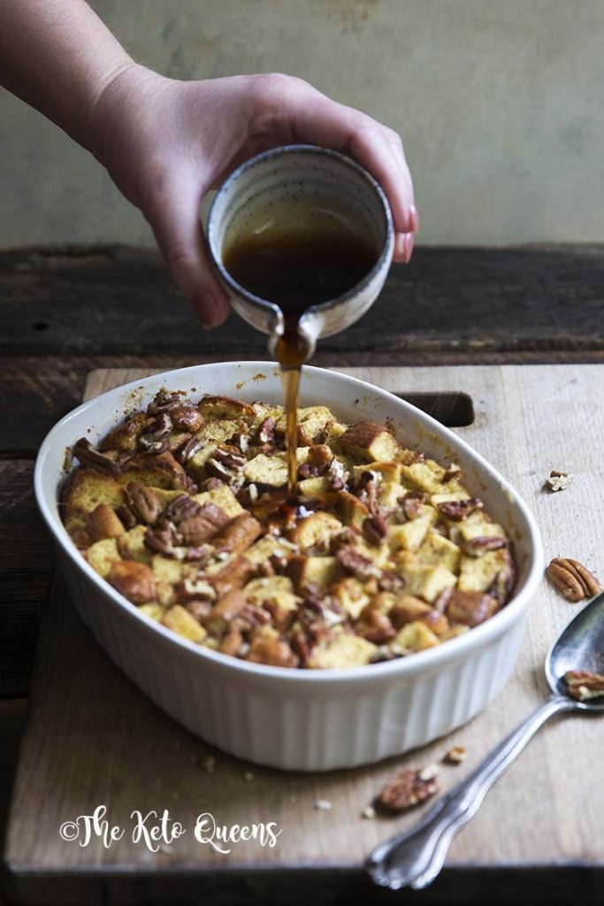 vertical image of maple pecan french toast casserole in a white casserole dish, with a spoon on a wood background with low carb syrup being poured onto it