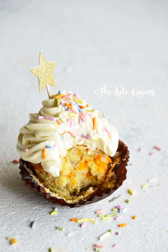 vertical image of a keto cupcake - vanilla cupcake with white chocolate buttercream frosting with sprinkles and a star with a bite taken out of it
